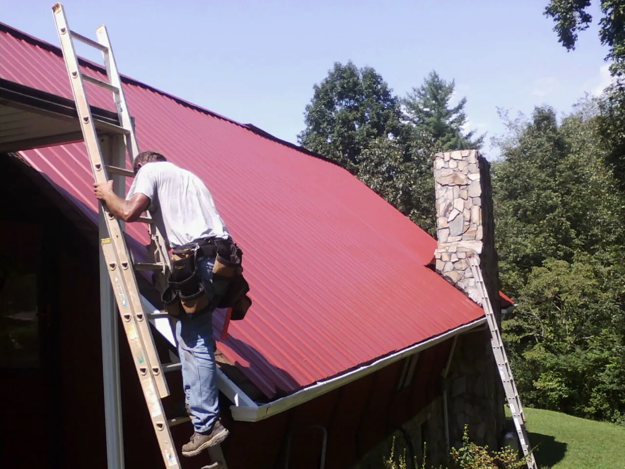 Roofer on a ladder beside a red metal roof and stone chimney — Soffit & Fascia Repair by Roofing Brothers in Derry